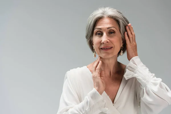 Happy middle aged bride in white wedding dress adjusting grey hair isolated on grey — Stock Photo