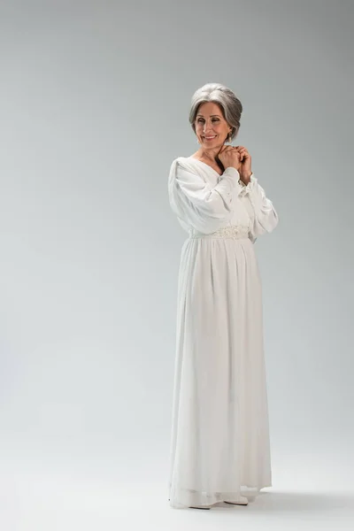 Повна довжина радісної нареченої середнього віку в білій весільній сукні, що стоїть на сірому — стокове фото