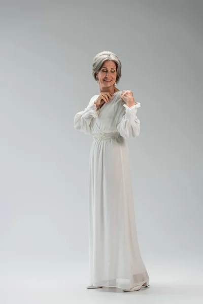 Full length of joyful middle aged bride adjusting white wedding dress on grey — Foto stock