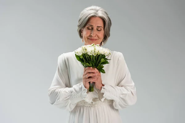 Joyful middle aged bride in white wedding dress holding bouquet and smelling flowers isolated on grey — Stock Photo