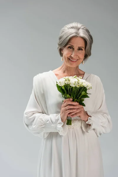 Cheerful middle aged bride in white wedding dress holding bouquet isolated on grey — Photo de stock