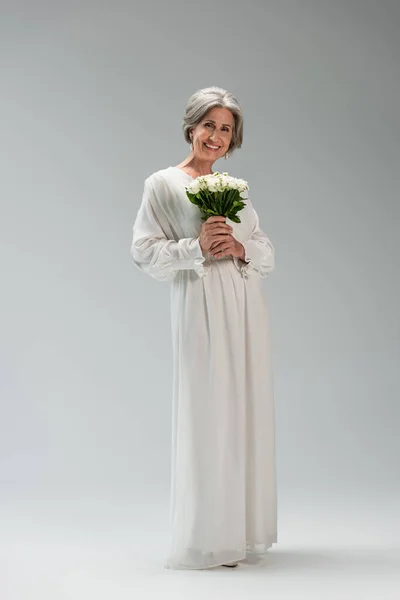 Full length of happy middle aged bride in white dress holding wedding bouquet on grey — Fotografia de Stock
