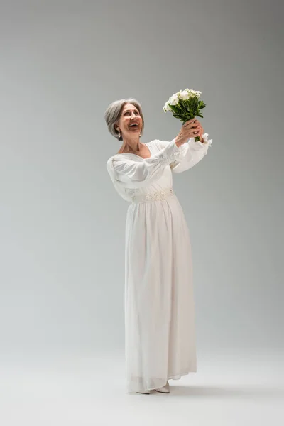 Full length of excited middle aged bride in white dress holding wedding bouquet on grey — Fotografia de Stock
