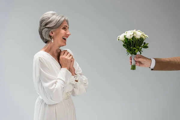 Groom holding wedding bouquet near happy middle aged bride in white dress isolated on grey — Stockfoto