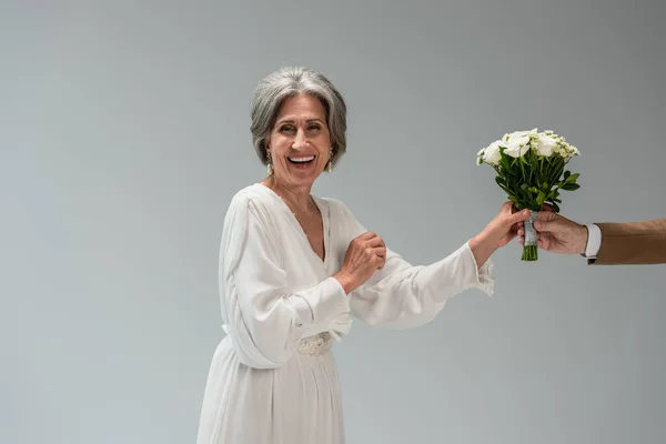 Groom giving wedding bouquet near joyful middle aged bride in white dress isolated on grey - foto de stock