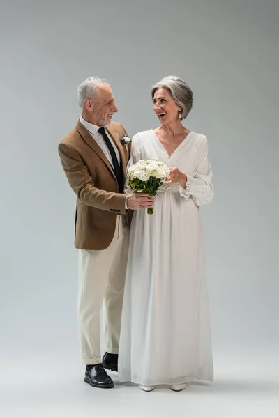 Full length of cheerful mature bride in wedding dress holding bouquet and standing with middle aged groom in suit on grey — Stockfoto