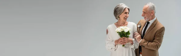 Happy bride in wedding dress holding bouquet and standing with middle aged groom in suit isolated on grey, banner — Stock Photo