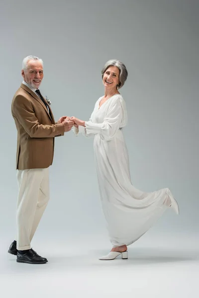 Full length of happy mature groom and bride smiling while holding hands on grey — Photo de stock