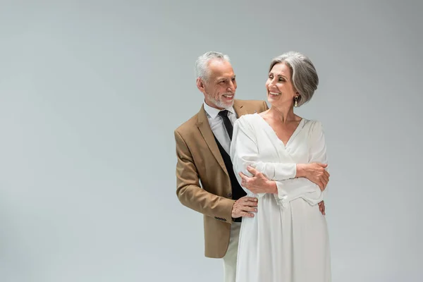 Happy middle aged groom hugging cheerful bride in wedding dress isolated on grey - foto de stock