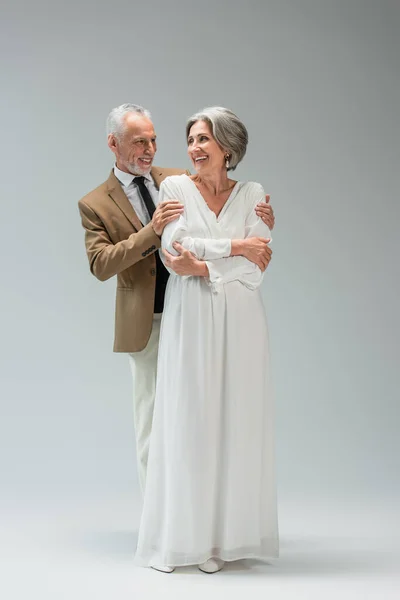 Full length of happy middle aged groom hugging cheerful bride in wedding dress on grey — Stock Photo