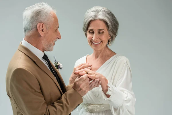 Middle aged groom wearing golden engagement ring on happy bride in white dress isolated on grey — Stock Photo