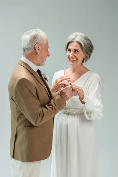 Зрелый мужчина в костюме с золотым обручальным кольцом на счастливой невесте в белом платье, изолированном на сером — стоковое фото