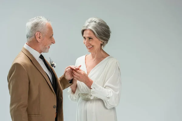 Happy mature bride in white dress wearing engagement ring on finger of middle aged groom isolated on grey - foto de stock