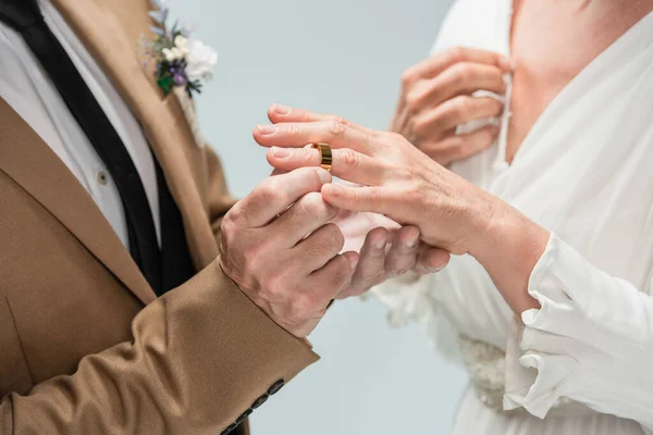 Cropped view of groom wearing golden engagement ring on finger of bride in white dress isolated on grey - foto de stock