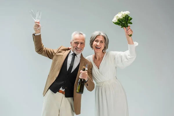 Cheerful middle aged groom holding bottle of champagne and glasses near happy bride in white dress isolated on grey — Stock Photo
