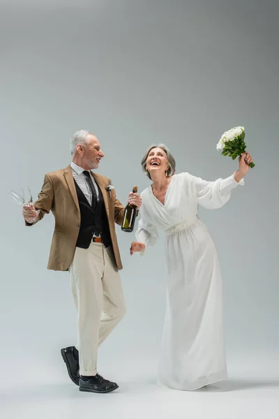 Full length of happy middle aged man holding bottle of champagne and glasses while dancing with bride in white dress on grey — Fotografia de Stock