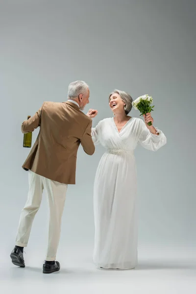 Full length of happy middle aged man holding bottle of champagne while dancing with cheerful bride in white dress on grey — Stock Photo