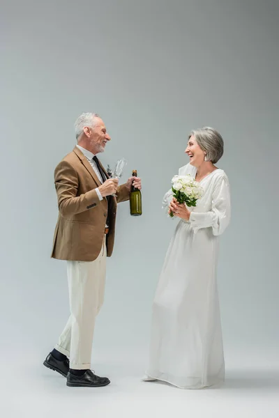 Full length of happy middle aged man holding bottle of champagne near cheerful bride in white dress on grey — Foto stock