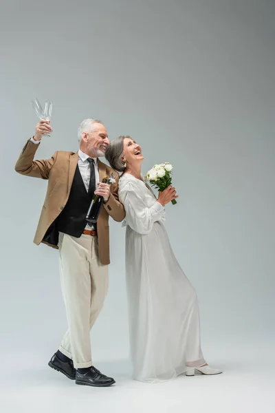 Full length of joyful middle aged man holding bottle of champagne and glasses near cheerful bride in white dress on grey — Photo de stock