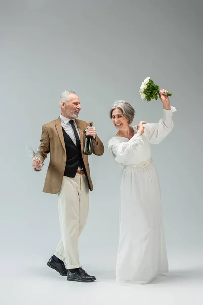 Full length of joyful middle aged man holding bottle of champagne and glasses and looking at cheerful bride on grey — Stockfoto