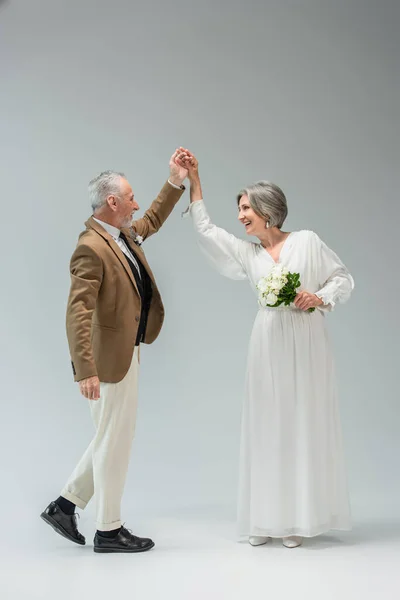 Full length of happy middle aged newlyweds dancing while holding hands on grey — Photo de stock