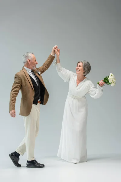 Full length of cheerful middle aged newlyweds dancing while holding hands on grey - foto de stock