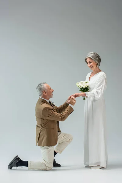 Full length of cheerful mature man standing on knee and wearing engagement ring on finger of bride with wedding bouquet on grey — Fotografia de Stock