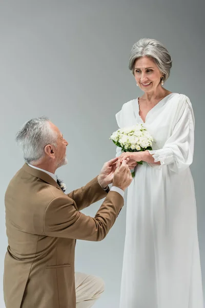 Mature man wearing engagement ring on finger of cheerful bride with wedding bouquet isolated on grey - foto de stock