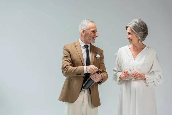 Happy middle aged groom opening bottle of champagne near woman in white dress holding glasses isolated on grey — Photo de stock