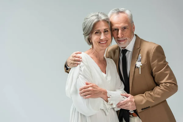 Cheerful middle aged man in suit with boutonniere hugging bride in wedding dress isolated on grey — Stockfoto