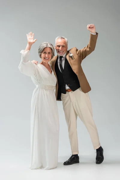 Full length of happy mature newlyweds waving hands standing smiling and standing on grey — Stockfoto