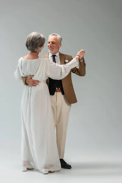 Повна довжина щасливого нареченого середнього віку і нареченої у весільній сукні тримає руки під час танців на сірому — стокове фото