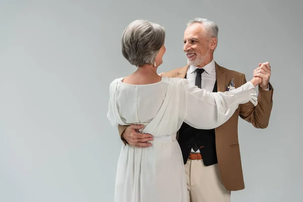 Smiling middle aged groom and bride in wedding dress holding hands while dancing isolated on grey - foto de stock