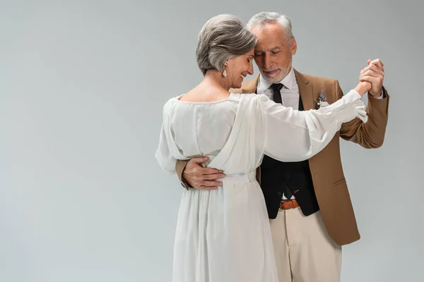 Pleased middle aged groom and happy bride in wedding dress holding hands while dancing isolated on grey — Stockfoto