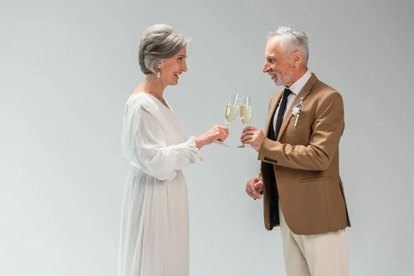 Side view of middle aged groom and happy bride in wedding dress clinking glasses of champagne isolated on grey — Fotografia de Stock