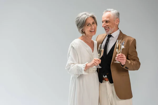 Middle aged groom and happy bride in wedding dress holding glasses of champagne isolated on grey — Stock Photo