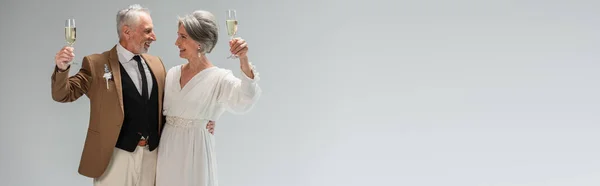 Жених среднего возраста и счастливая невеста в белом платье с бокалами шампанского изолированы на сером, баннер — стоковое фото