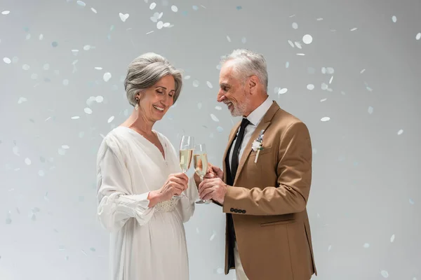 Middle aged groom and happy bride in white dress clinking glasses of champagne near falling confetti on grey — Photo de stock