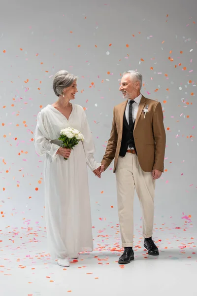 Full length of cheerful middle aged groom and happy bride in white dress holding hands under falling confetti on grey — Fotografia de Stock