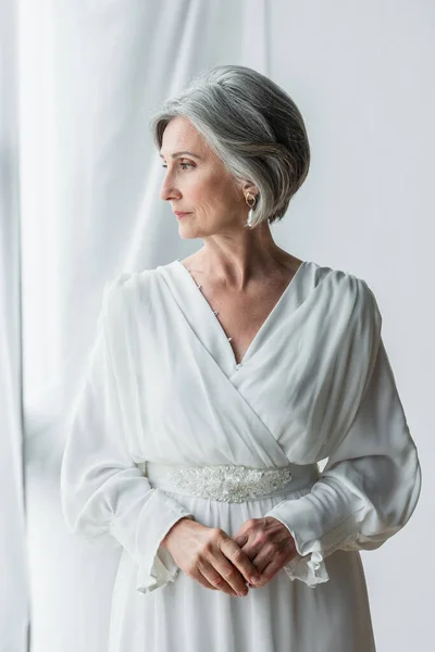 Elegant and middle aged bride in white dress standing near curtains and window — Foto stock