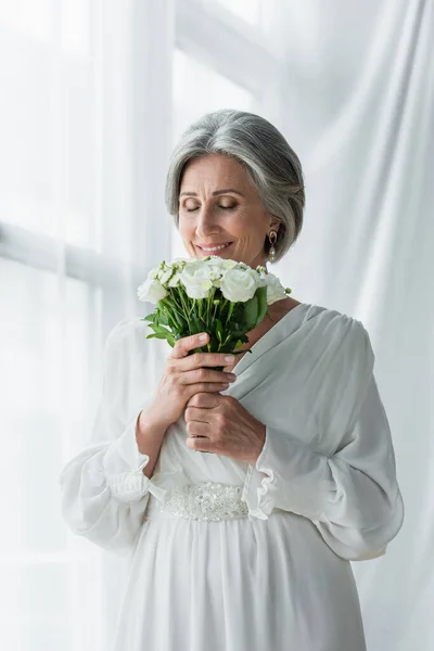 Happy middle aged bride in white dress smelling wedding bouquet near curtains — Stock Photo