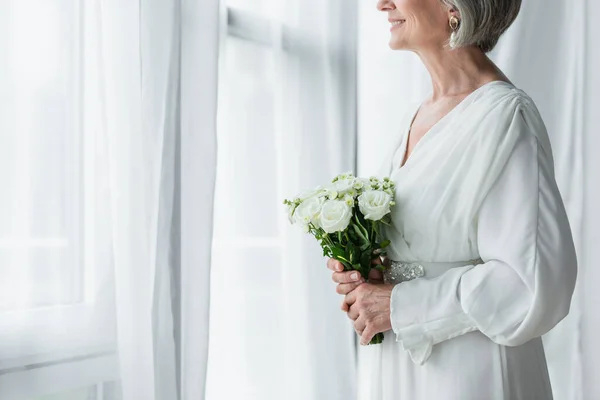 Обрізаний вид щасливої нареченої середнього віку в білій сукні, що тримає весільний букет і стоїть біля штор — стокове фото