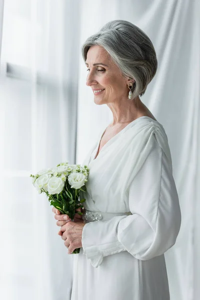 Pleased middle aged woman in white dress holding wedding bouquet and standing near curtains - foto de stock