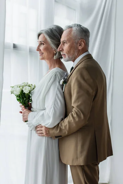Бородатий середнього віку наречений обіймає щасливу наречену в білій сукні з весільним букетом — стокове фото