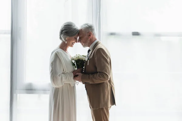 Side view of bearded middle aged groom holding wedding bouquet with happy bride in white dress — Stockfoto