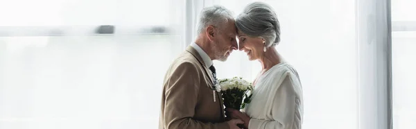 Side view of bearded middle aged groom holding wedding bouquet with happy bride in white dress, banner — Foto stock