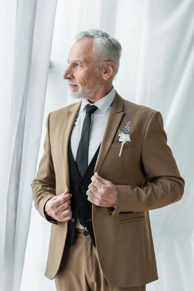 Bearded middle aged man in suit with boutonniere posing near white curtains - foto de stock