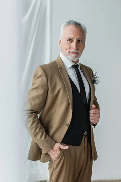 Bearded middle aged man in suit with boutonniere smiling while posing near white curtains — Foto stock