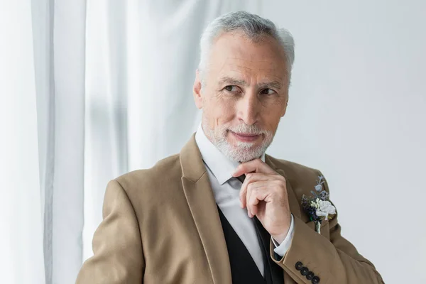 Bearded middle aged man in suit with floral boutonniere smiling while looking away near white curtains - foto de stock