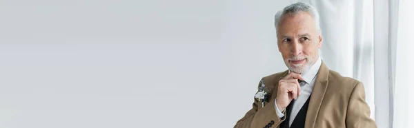 Бородатый мужчина средних лет в костюме с цветочным бутоньером улыбается, глядя в сторону белых занавесок, баннер — стоковое фото